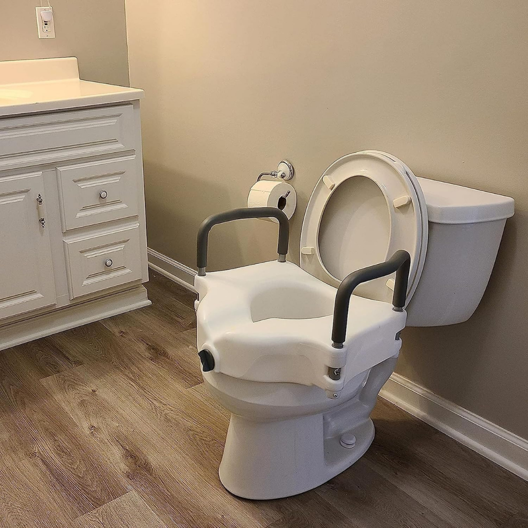 A bathroom features a white toilet with the ProBasics E-Z Lock 4.5 Raised Toilet Seat with Handles, next to a white vanity and wall-mounted toilet paper holder. The floor has wood-patterned laminate.