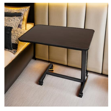 The Roscoe Rolling Overbed Table, a compact black non-tilting bedside table with wheels, stands beside a bed with a beige upholstered headboard and patterned pillows on a light-colored floor.
