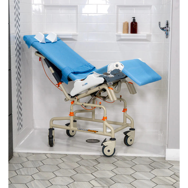 A Rifton Wave Pediatric Shower Chair with blue padding and wheels is placed in a white-tiled shower. Shampoo and soap bottles are on the shelf, and the floor has a geometric tile pattern.