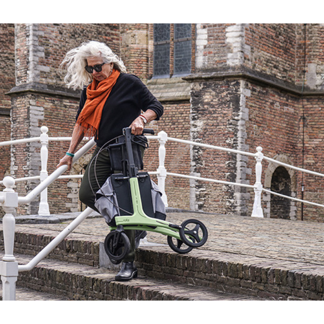 An older woman with white hair, sunglasses, a black top, and an orange scarf carries the Rollz Air Lightweight Folding Carbon Rollator (10.4 lbs) down outdoor brick steps beside a railing and historic brick building.