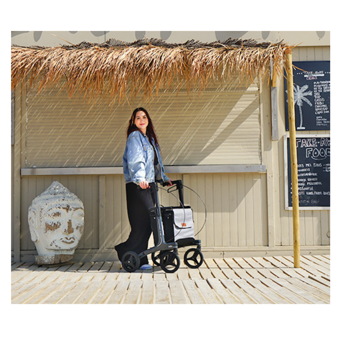 A woman in a denim jacket uses the Rollz Air Lightweight Folding Carbon Rollator (10.4 lbs) on a wooden deck near a thatched-roof structure, with a Buddha head and menu board in the background.