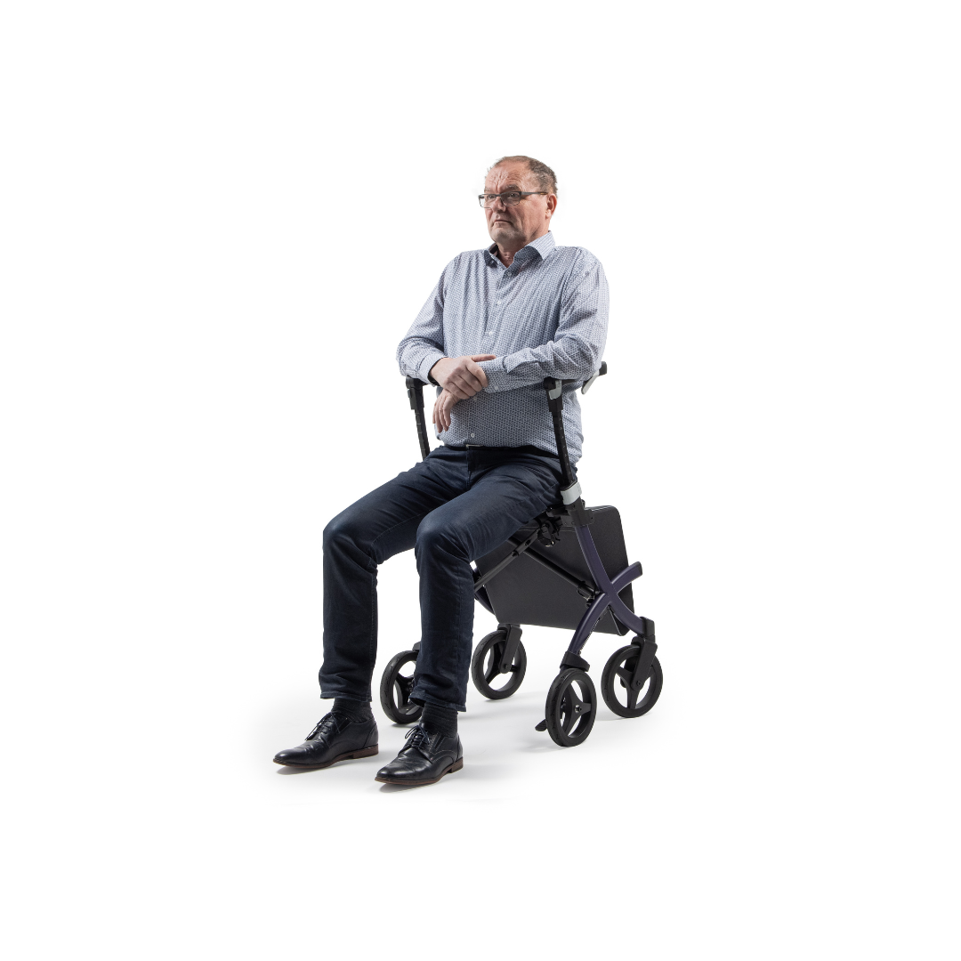 A man in glasses and a blue shirt is seated, facing forward, on a Rollz FLEX 2 Lightweight Rollator Walker featuring four wheels, a compact design, and an XL storage bag. The white background highlights this mobility aid.