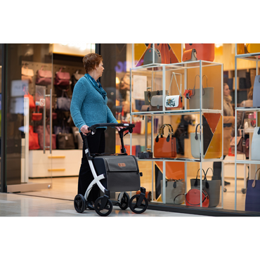 A woman with short hair uses a Rollz FLEX 2 Lightweight Rollator Walker featuring an XL storage bag and seat to browse a store display of colorful handbags. Shes dressed in a blue sweater and plaid pants amid the vibrant, stylish shop window.