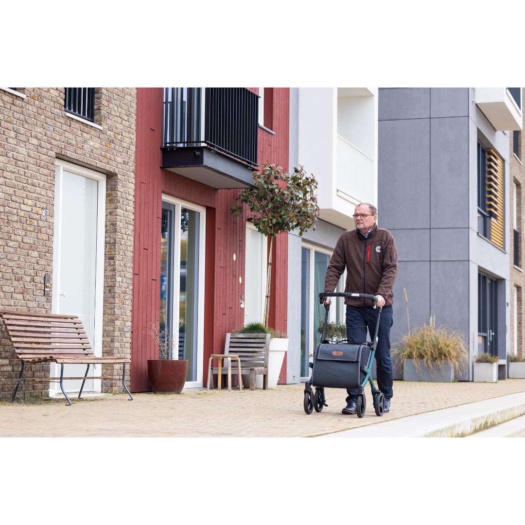 A man strolls with his Rollz FLEX 2 Lightweight Rollator Walker with XL Storage Bag & Seat along a modern residential building featuring colorful facades. Nearby, a wooden bench and potted plants by the entrance create a calm suburban vibe.