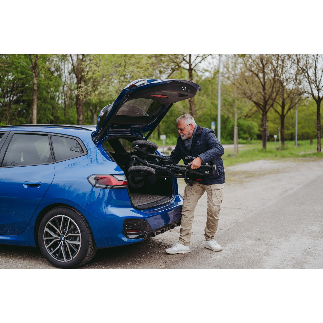 A man wearing a black jacket and beige pants loads a folded Rollz Motion Electric 3-in-1 Rollator, Transport Chair & Powerchair into the open trunk of a blue car while fresh green-leaved trees stand in the park background.