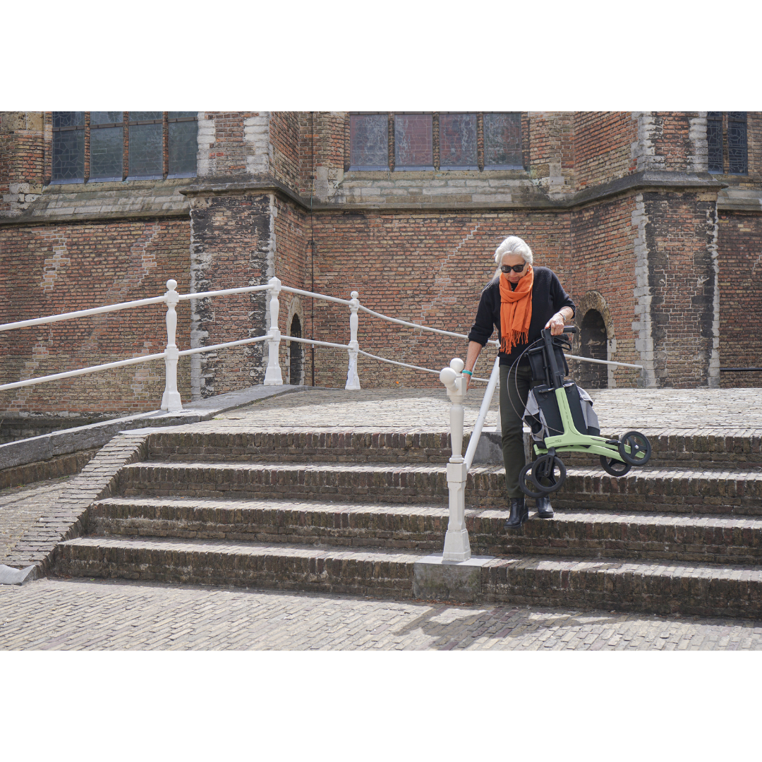 An older woman with white hair, sunglasses, and an orange scarf carefully descends stone steps outside a historic brick building, holding the handrail while using her Rollz Air Lightweight Folding Carbon Rollator, which weighs just 10.4 lbs.
