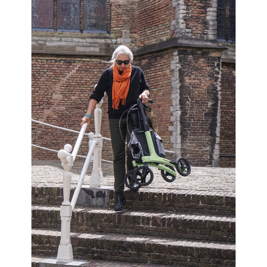 An older woman with gray hair, sunglasses, and an orange scarf walks down outdoor steps beside a brick building, holding a green trolley and using the Rollz Air Lightweight Folding Carbon Rollator, which weighs just 10.4 lbs.