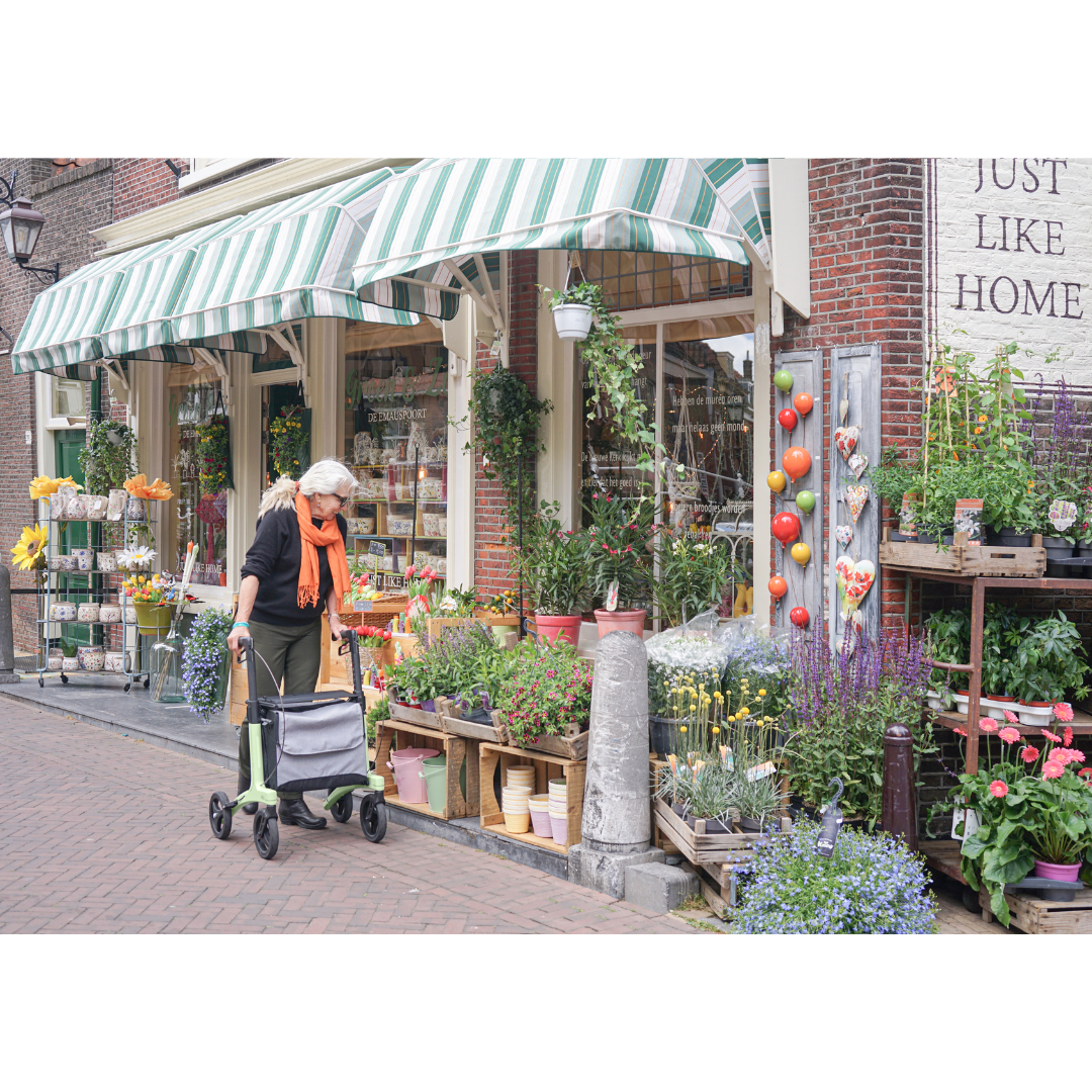 An older woman with a Rollz Air Lightweight Folding Carbon Rollator (10.4 lbs) passes a flower shop with striped green awnings and colorful potted plants outside. A sign on the brick wall reads “Just Like Home.”.