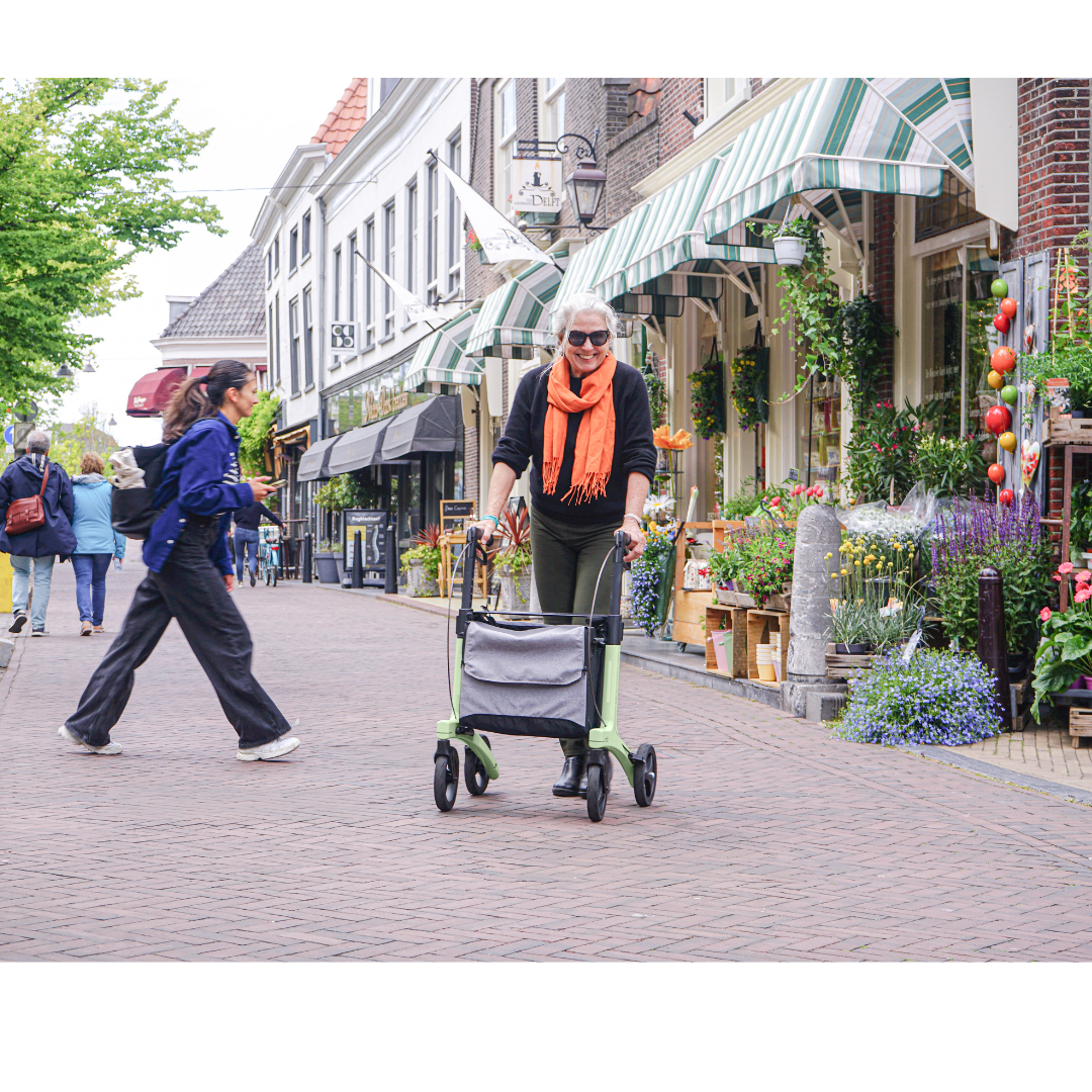 An older woman with white hair and an orange scarf uses the Rollz Air Lightweight Folding Carbon Rollator (10.4 lbs) on a cobblestone street lined with shops and flowers, while another person crosses ahead. The lively scene bursts with vibrant colors.