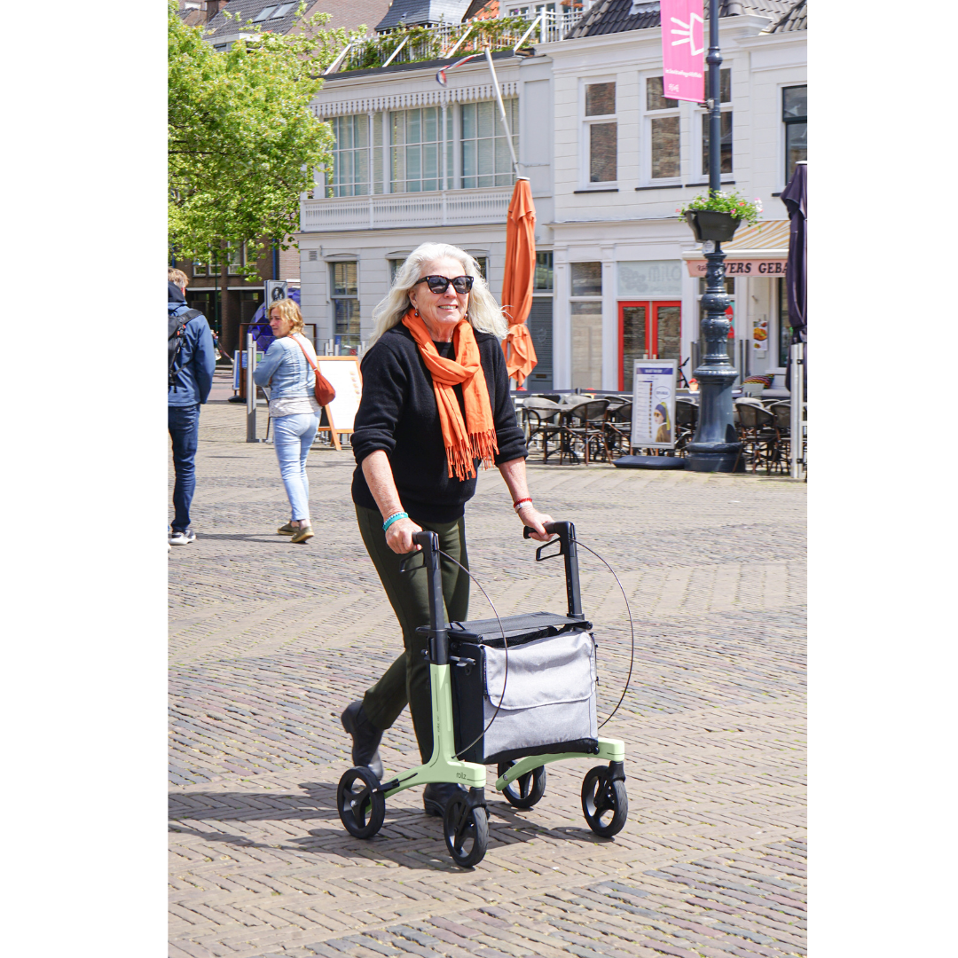 An older woman with long gray hair and an orange scarf uses the Rollz Air Lightweight Folding Carbon Rollator (10.4 lbs) as she strolls through a sunny town square with cobblestone streets, buildings, and people in the background.