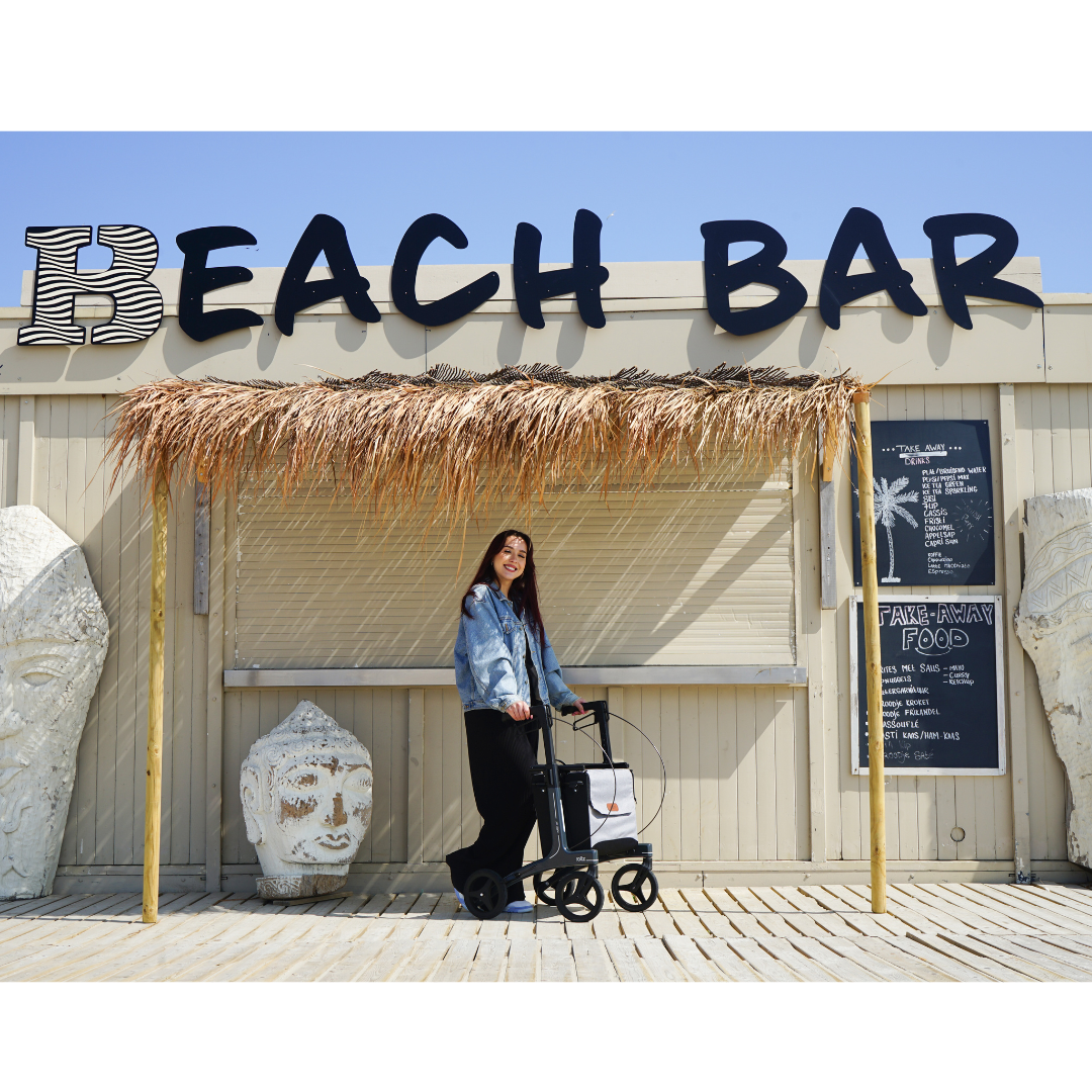 A woman smiles with her stroller and the Rollz Air Lightweight Folding Carbon Rollator (weighs 10.4 lbs) under a thatched canopy by a building marked BEACH BAR, with two large decorative heads and a blackboard menu at the entrance.