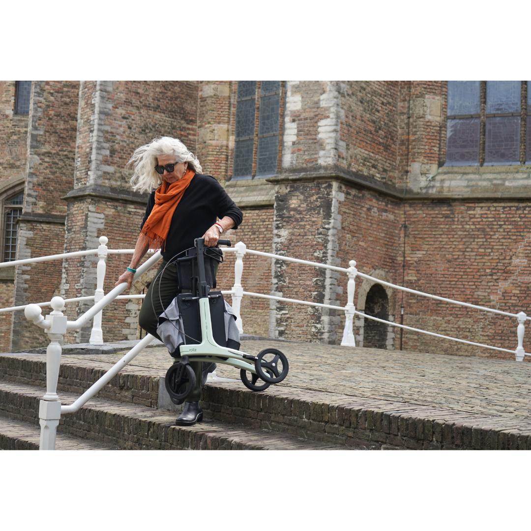 A woman with white hair, sunglasses, and an orange scarf pulls the Rollz Air Lightweight Folding Carbon Rollator (10.4 lbs) down stone steps beside a historic brick building.