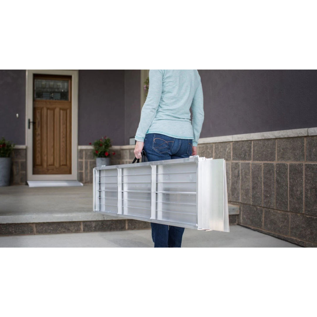 Wearing jeans and a light blue sweater, a person carries an EZ-Access SUITCASE® Singlefold Mobility Ramp—portable and folded—outside a house with a wooden door and stone porch.