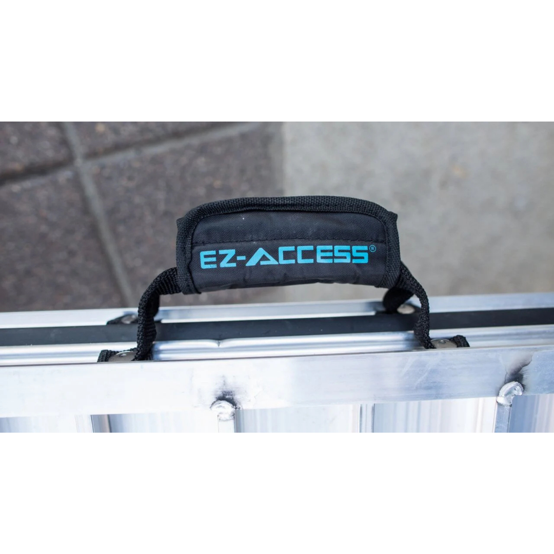 A close-up of a black handle with the blue EZ-ACCESS logo attached to the silver EZ-Access SUITCASE® Singlefold Mobility Ramp - Portable, with a blurred stone or concrete ground in the background. See Specifications and our Videos for more details.