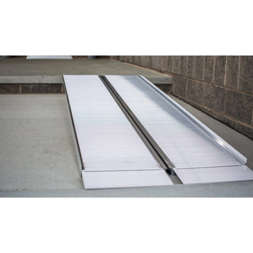 The EZ-Access SUITCASE® Singlefold Mobility Ramp - Portable is set up over a curb by concrete steps and a brick wall, providing an accessible path from the sidewalk to the entryway. Check specifications and warranty details before use.