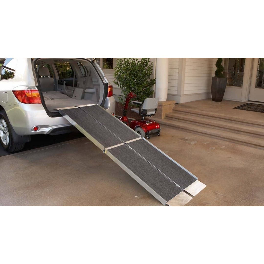 An EZ-Access SUITCASE® TRIFOLD® Portable AS Ramp extends from the back of a car to the ground, providing easy vehicle access. A red mobility scooter is parked nearby in front of a house with steps and a potted plant.