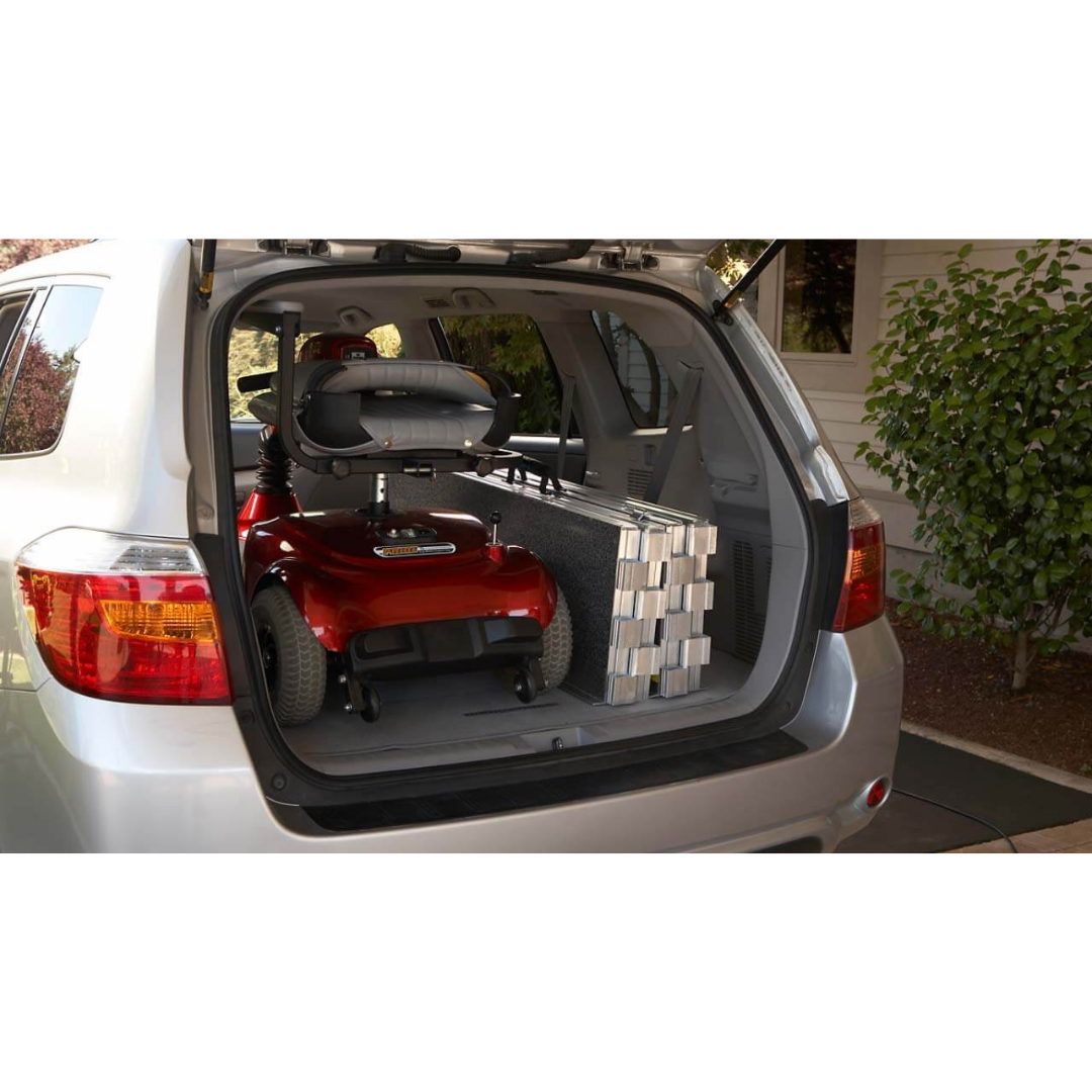 The open SUV trunk displays an EZ-Access SUITCASE® TRIFOLD® Portable AS Ramp folded next to a red mobility scooter, with greenery and a porch seen in the background.