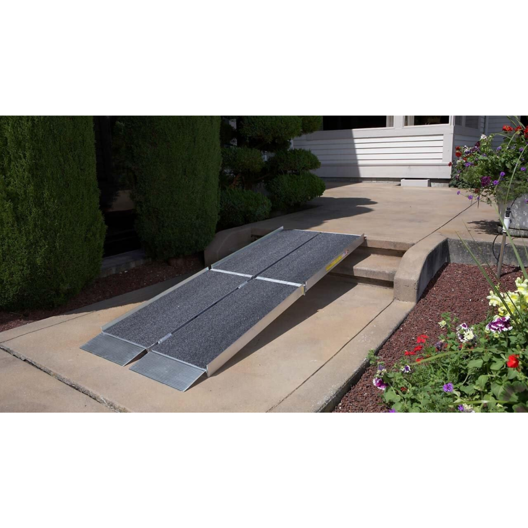 The EZ-Access SUITCASE® TRIFOLD® Portable AS Ramp, made of aircraft-grade aluminum, spans a curb and step to connect the garden walkway with the house entrance surrounded by shrubs and flowers.