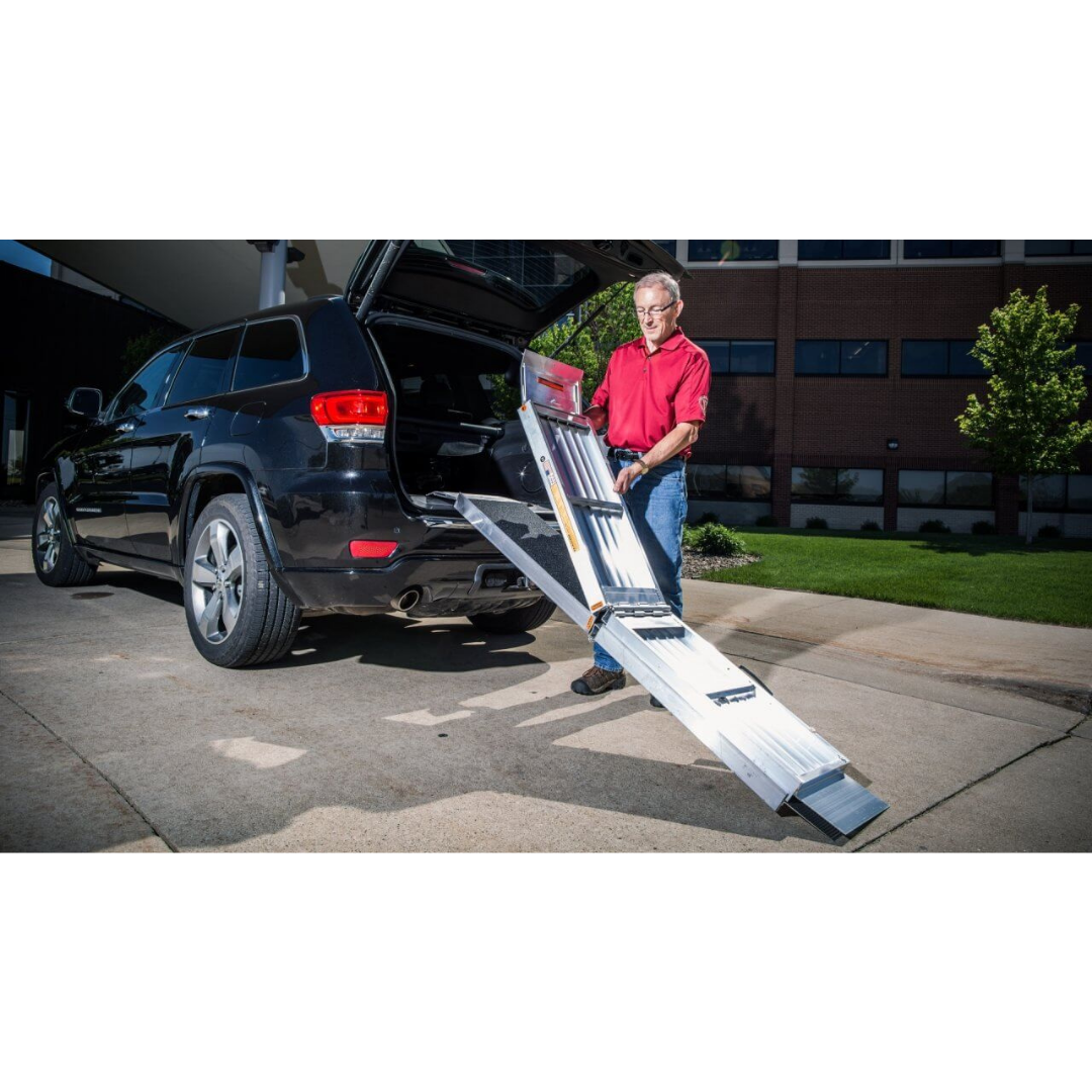 A person in a red shirt sets up the EZ-Access SUITCASE® TRIFOLD® Portable AS Ramp from an SUVs back outdoors, ready to load or unload, with a modern building and landscaped greenery in the background.