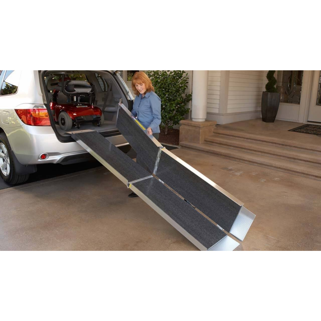 A woman sets up an EZ-Access SUITCASE® TRIFOLD® Portable AS Ramp with a slip-resistant surface from her SUV, preparing to unload a red mobility scooter in front of a house with steps.