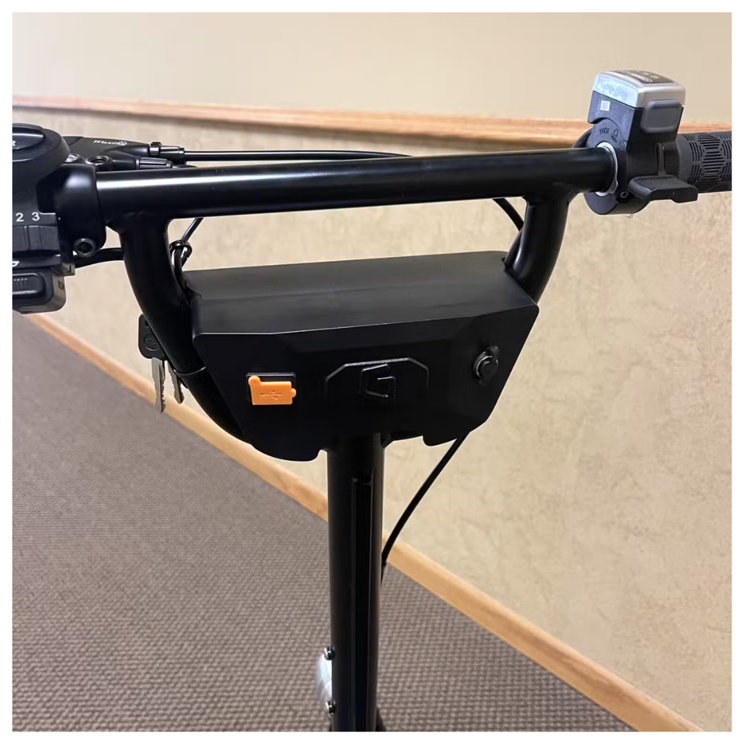 The close-up of Glion SNAPnGO Scooter Accessories displays a key slot, orange switch, and mounted control box on the handlebars. A textured grip and digital display showcase efficient design against a neutral indoor background.