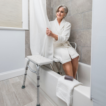 An older woman sits on the Drive Splash Defense™ Transfer Bench with Curtain Guard Protection in a bathtub, holding the shower curtain. A towel and soap rest nearby. The bathroom has gray tiles and wooden flooring for added bath safety.