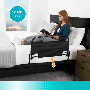 A woman sits upright in bed reading, with the Stander Safety Bed Rail & Padded Pouch—30" rail length—attached for fall protection. An orange arrow highlights the bed’s edge in a modern, minimal room.