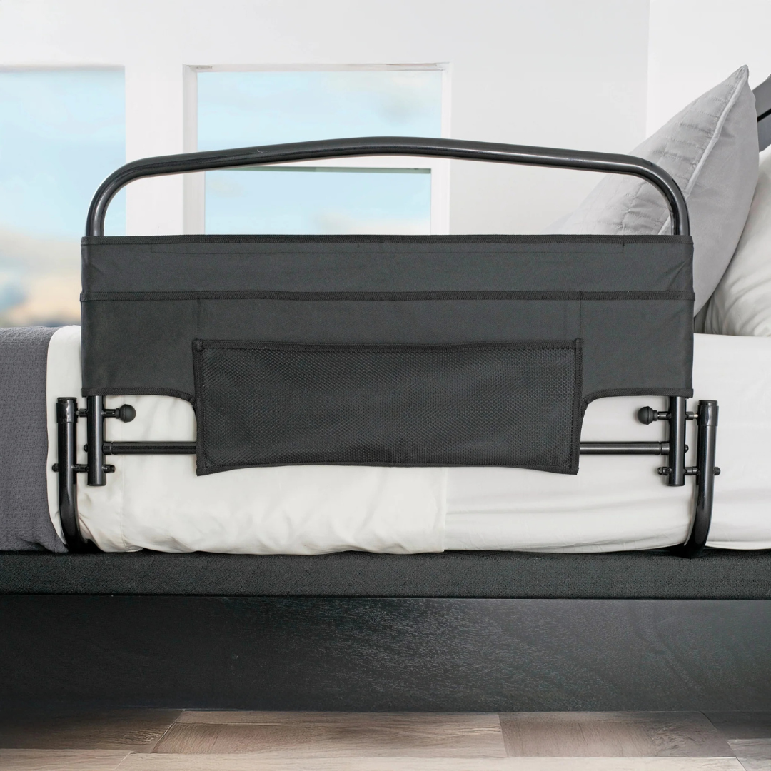 The Stander Safety Bed Rail & Padded Pouch (30" rail) in black features a mesh storage pocket and attaches securely between the mattress and bed frame, providing fall protection in a modern bedroom with bright windows.