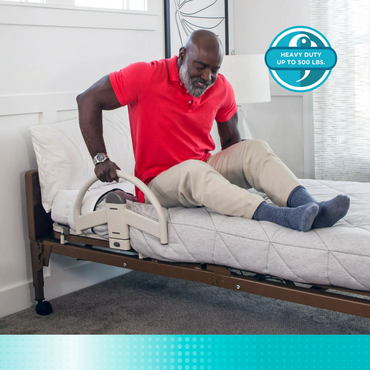 A man in a red shirt uses the Stander EZ Click LTC Bariatric Bed Handle (Set of 2) for support while sitting on a bed. A circular label states: “Heavy Duty Up to 500 lbs.”.