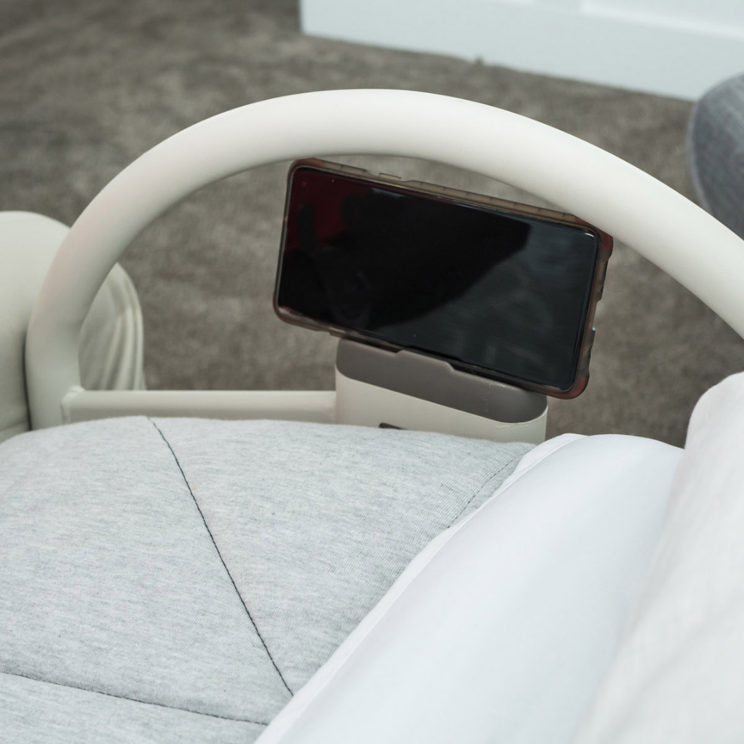 A smartphone is mounted on the white Stander EZ Click LTC Bariatric Bed Handle (set of 2) attached to a hospital bed rail beside a gray cushioned seat, with soft gray carpet in the background.