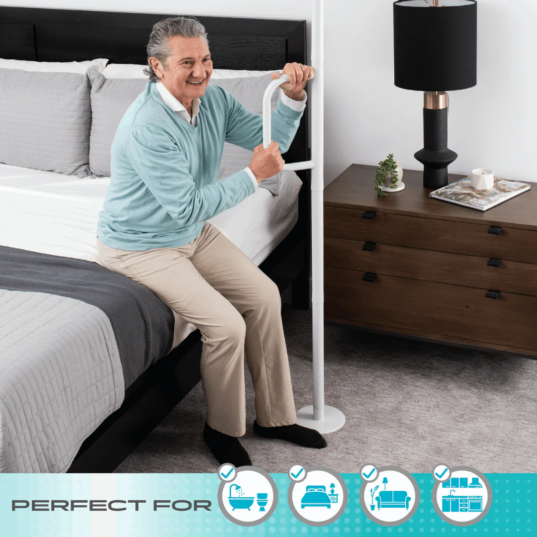 An older adult smiles while holding the Stander Wonder Pole Lite—an adjustable, tension-mounted safety pole for beds, chairs, and bathrooms. The bedroom features a nightstand, lamp, and gray bedding. Ideal for fall prevention at home.