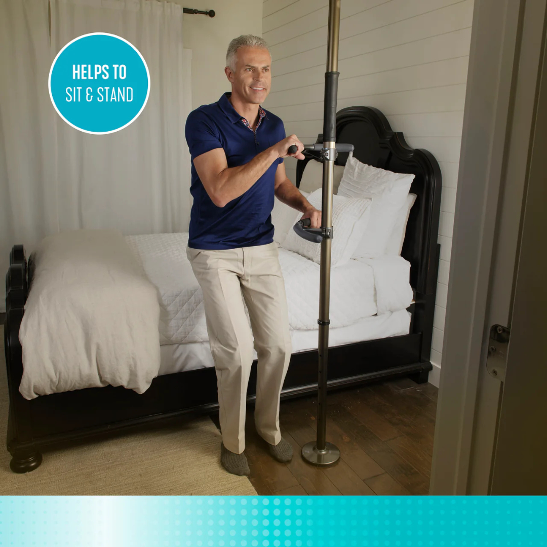 A smiling older man uses the Stander Wonder Pole Premium—Fall Prevention Pole, tension-mounted beside a bed, to help himself stand. Text reads, Helps to sit & stand. The bedroom has a dark bed frame, white bedding, and wood floors.