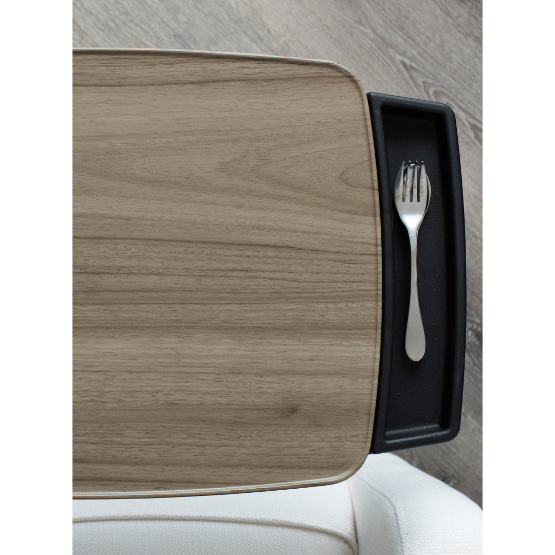 The Stander Wonder Tray Premium - XL TV Tray & Standing Aid is pictured from above, with a metal fork in its black tray, beside a rectangular light wood tabletop. A white chair and light wood floor are also visible.