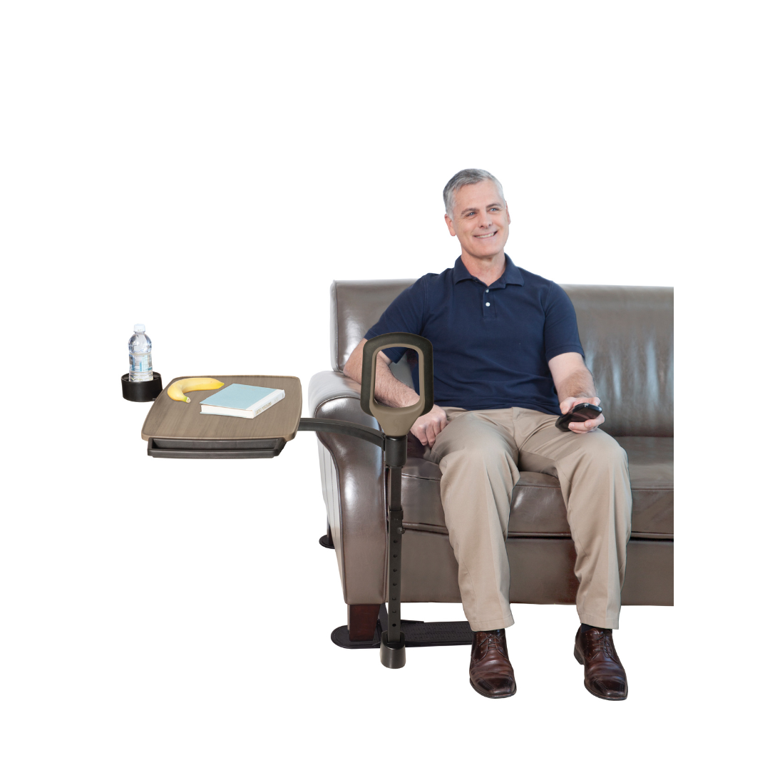 A smiling man sits on a brown leather sofa, holding a remote. Beside him is the Stander Wonder Tray Premium—an XL TV tray with cup holder, water bottle, and book, attached to a sturdy standing aid handle for convenience.