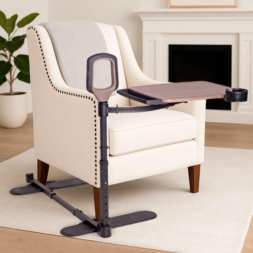 The Stander Wonder Tray Premium - XL TV Tray & Standing Aid, a beige armchair accessory with dark-brown adjustable tray, stand assist handle, and cup holder, sits in a modern living room with a plant and fireplace in the background.