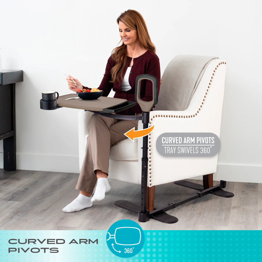 A woman enjoys a meal from the Stander Wonder Tray Premium—an XL TV tray with a swivel 360-degree tray, curved support arm, stand assist handle for safer dining, and space for her coffee mug—while seated in a white armchair.
