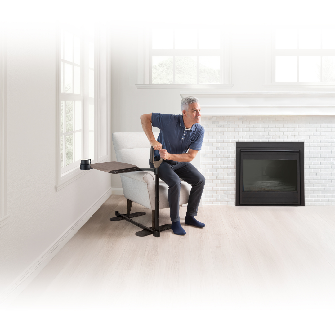 An older man sits by a fireplace in a bright room, using the Stander Wonder Tray Premium—an XL TV tray and standing aid—to hold his mug while relaxing in his chair.