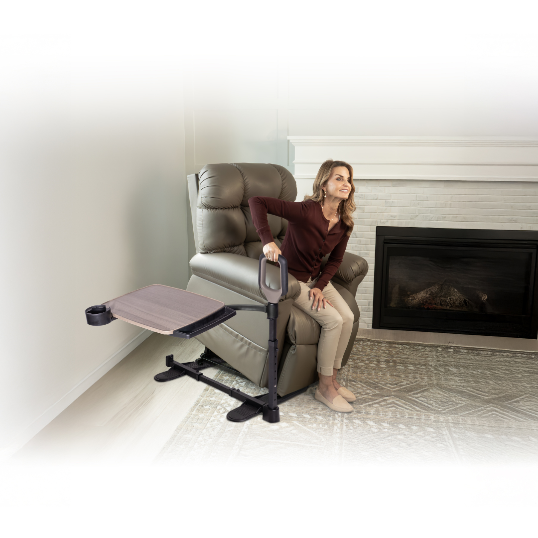 A woman uses the Stander Wonder Tray Premium - XL TV Tray & Standing Aid, attached to her lift chair, to help herself stand. The adjustable tray with a cup holder is also connected. The scene is in a living room with fireplace and patterned rug.