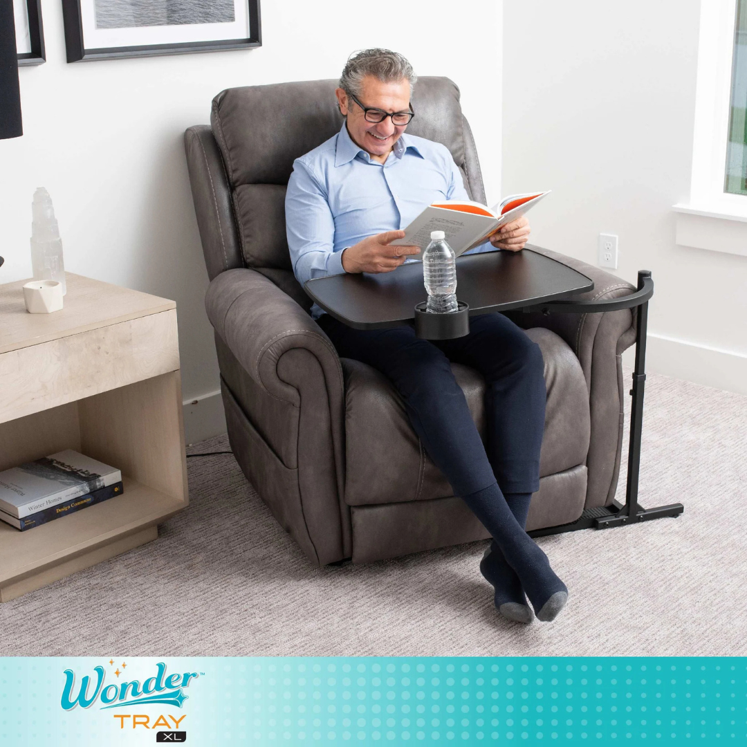 A man relaxes in a gray recliner, reading and smiling. The Stander Wonder Tray XL - Swivel Living Room Tray Table, featuring a cup holder with a water bottle, sits over his lap. The bright room has a modern style.