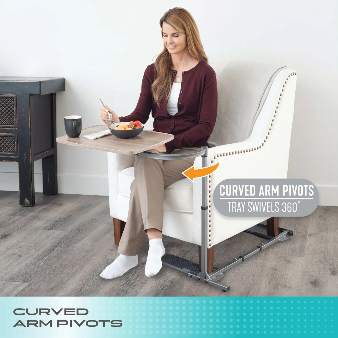 A woman sits in a white armchair using the Stander Wonder Tray—an adjustable, 360° swivel living room tray table with a metal support, attached to the armrest, holding food and a drink. It’s also ideal as a laptop stand.