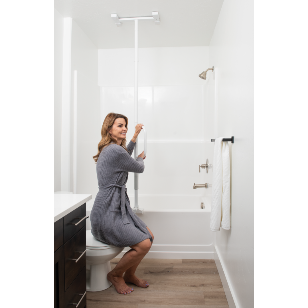 A woman in a gray robe sits on a toilet in a white bathroom, holding the Stander Wonder Bathtub Pole Lite for support. Towels hang on a nearby rack, and the pole is installed vertically between the floor and ceiling.