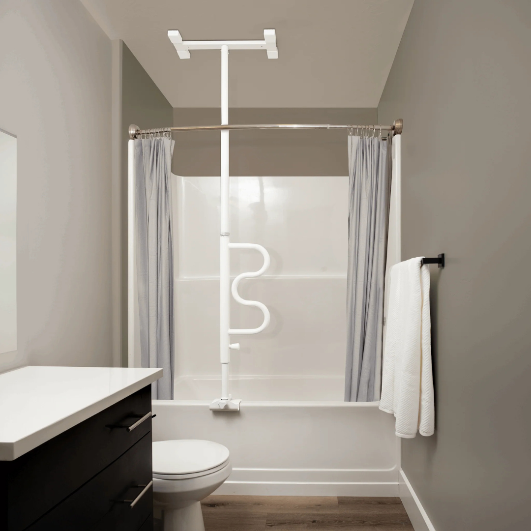 A modern bathroom with a Stander Wonder Bathtub Pole for safety next to a white tub and shower, gray curtains, a towel on a black rack, white countertop, dark drawers, light gray walls, and wood flooring.