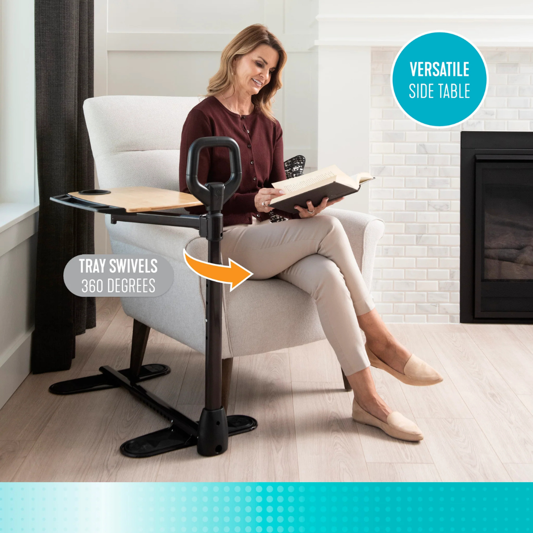 A woman sits reading in a light armchair, using the Stander Assist-A-Tray—an adjustable standing aid with a black 360° swivel tray and handle—positioned over her lap. A fireplace and window are visible in the background.