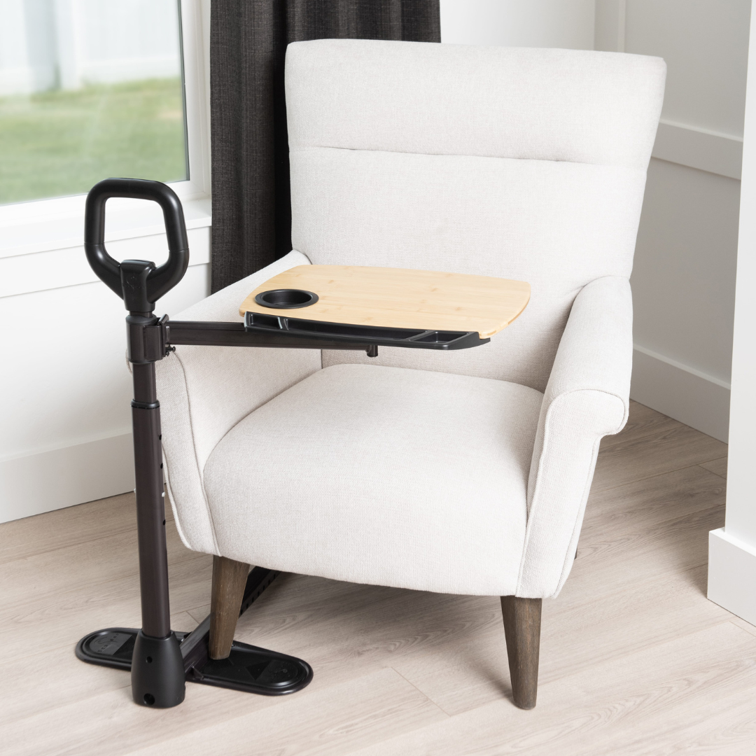 A light beige armchair with wooden legs sits by a window. In front is the Stander Assist-A-Tray, a standing aid with a stable handle, base, adjustable swivel tray, and cup holder for mobility and convenience.
