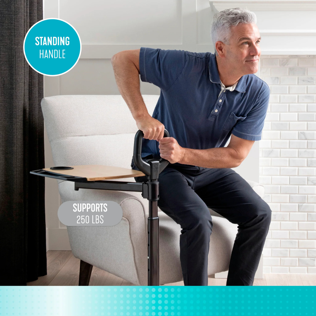 A man uses the Stander Assist-A-Tray Standing Aid With Swivel Tray, featuring a support handle and adjustable tray with cup holder, to help him stand up from an armchair. The handle supports up to 250 lbs.