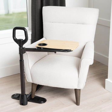 The Stander Assist-A-Tray, a light gray armchair with a wooden swivel tray and black standing handle, sits on a light wood floor by a window and dark curtain.