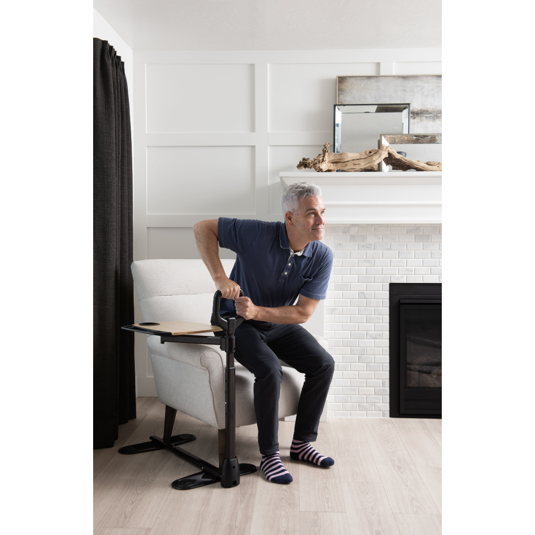 A gray-haired man sits indoors, gripping the Stander Assist-A-Tray—Standing Aid With Swivel Tray—to help himself stand up from his armchair. He wears striped socks; a fireplace and mirror are visible in the background.