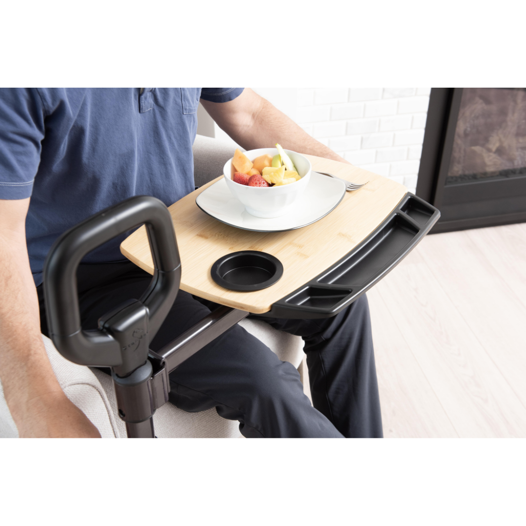 A person uses the Stander Assist-A-Tray—Standing Aid With Swivel Tray—attached to a mobility aid handle, sitting in a living room with light wood flooring. The tray holds a bowl of fruit, fork, has a cup holder, and provides convenient access.