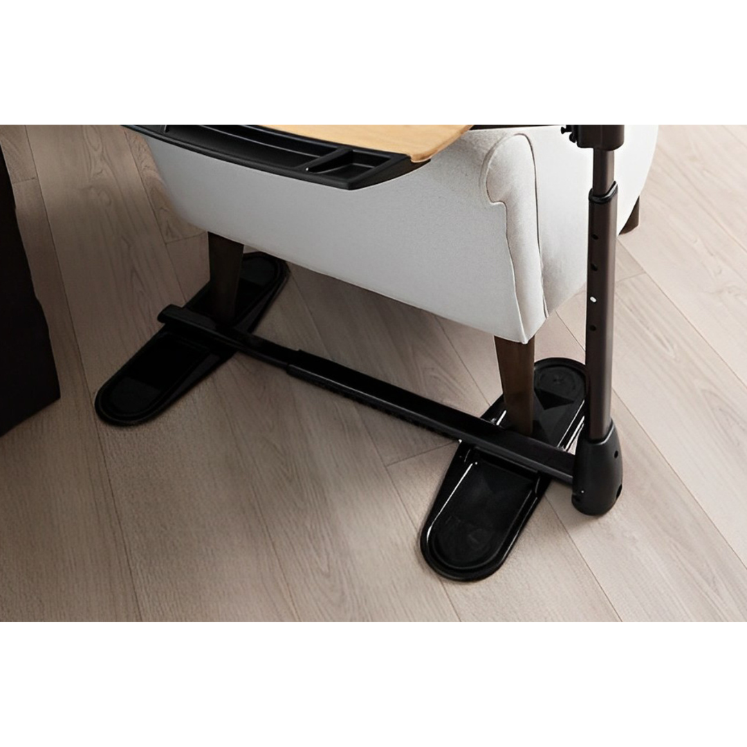 A close-up of the Stander Assist-A-Tray Standing Aid with footrests and a standing handle positioned under a light-colored armchair on a wooden floor.