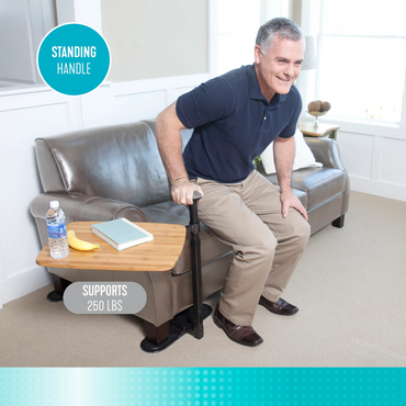 A smiling older man uses the Stander Omni Tray – TV Tray & Standing Aid, attached to a wooden table, to stand up from a couch. The tray holds a water bottle, banana, and book. Text: “STANDING HANDLE” and “SUPPORTS 250 LBS.”.